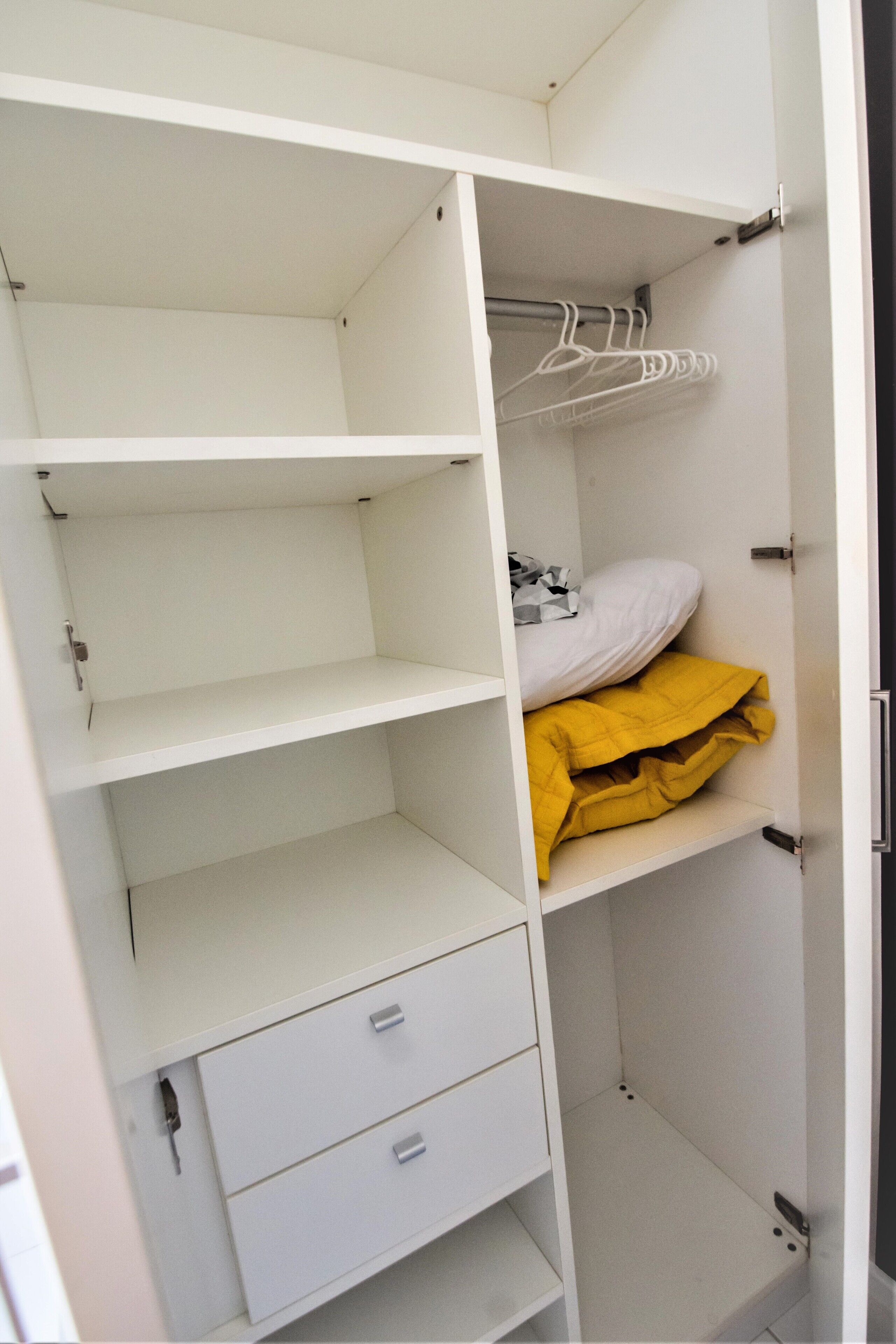 Un armario blanco con puertas abiertas, mostrando estantes, una barra para colgar ropa y una manta amarilla doblada.