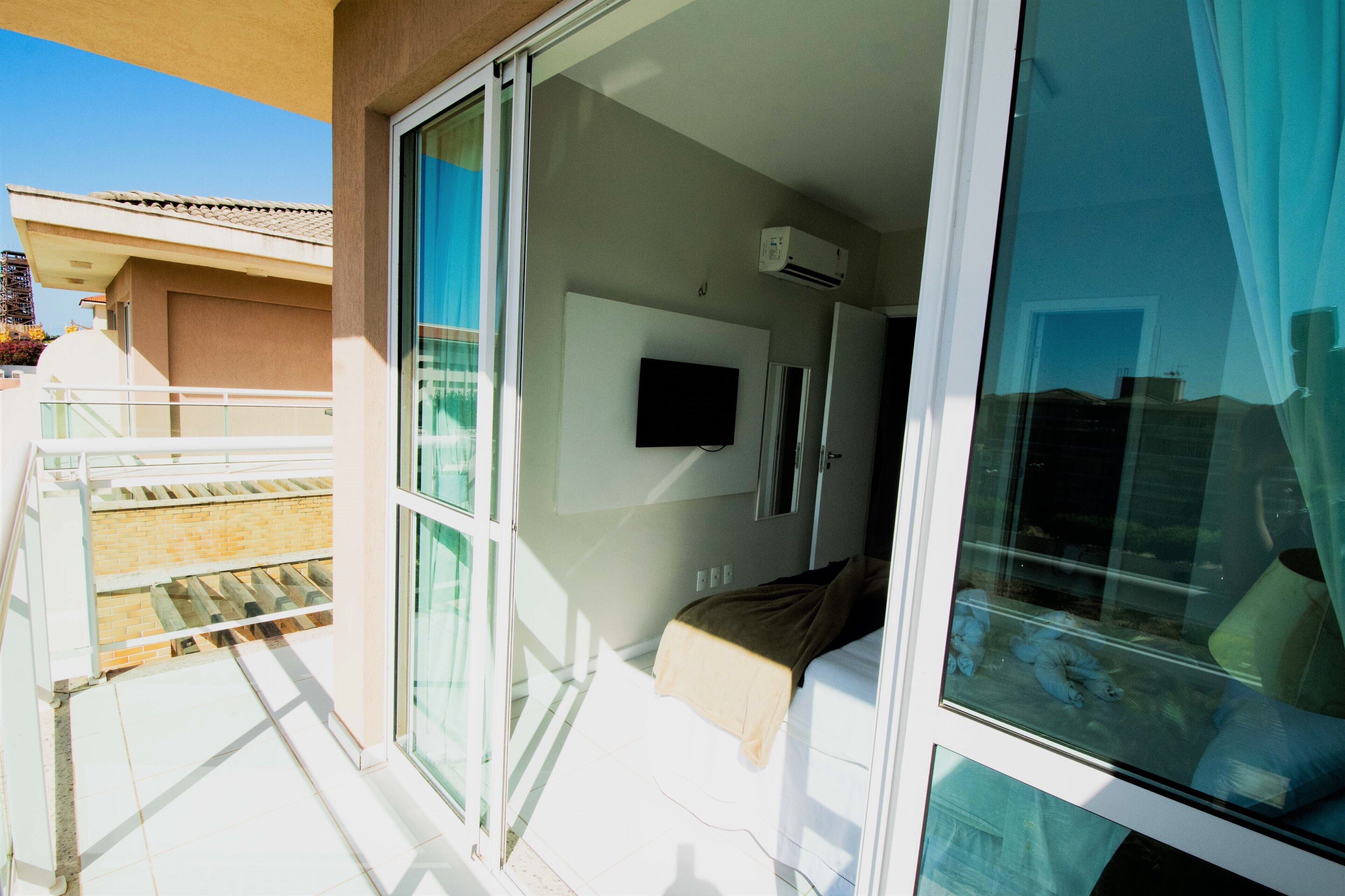 A hotel room with a sliding glass door leading to a balcony, a bed with a pillow, a mounted TV, and an air conditioning unit.