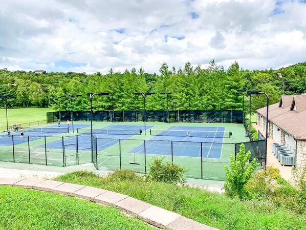 A tennis court with multiple courts, surrounded by a fence, and a building with a stone facade in the background.