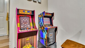 Zwei Spielautomaten, einer mit der Aufschrift 'Pac-Man' und der andere mit 'Mortal Kombat', in einem Raum mit Holzboden und einer weißen Tür.