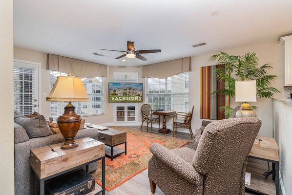 A living room with a sofa, coffee table, lamp, and a ceiling fan. There is a dining area with chairs and a table. A large painting is on the wall, and a plant is on the right side. - No Stairs! Condo Minutes from Branson Strip! (Branson)