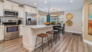 Una cocina moderna con armarios blancos, electrodomésticos de acero inoxidable, encimera de mármol y una zona con taburetes de bar.