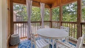 Un espace extérieur abrité, doté d’une table ronde et de chaises, d’une terrasse en bois et offrant une vue sur des arbres et un bâtiment en arrière-plan.