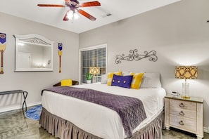 A bedroom with a bed, a ceiling fan, a mirror, a window with blinds, a nightstand with a lamp, and a decorative wall piece.