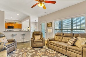A spacious living room with a brown leather sofa, a recliner, and a patterned rug. A ceiling fan is visible above.
