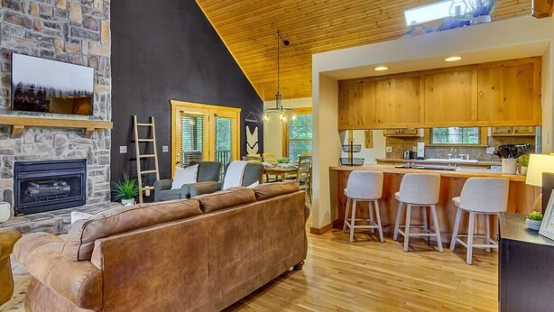 A modern living room with a stone fireplace, a large brown sofa, and a kitchen area with wooden cabinets and bar stools. - Modern Lodge, 3 Miles from Silver Dollar City! (Reeds Spring)