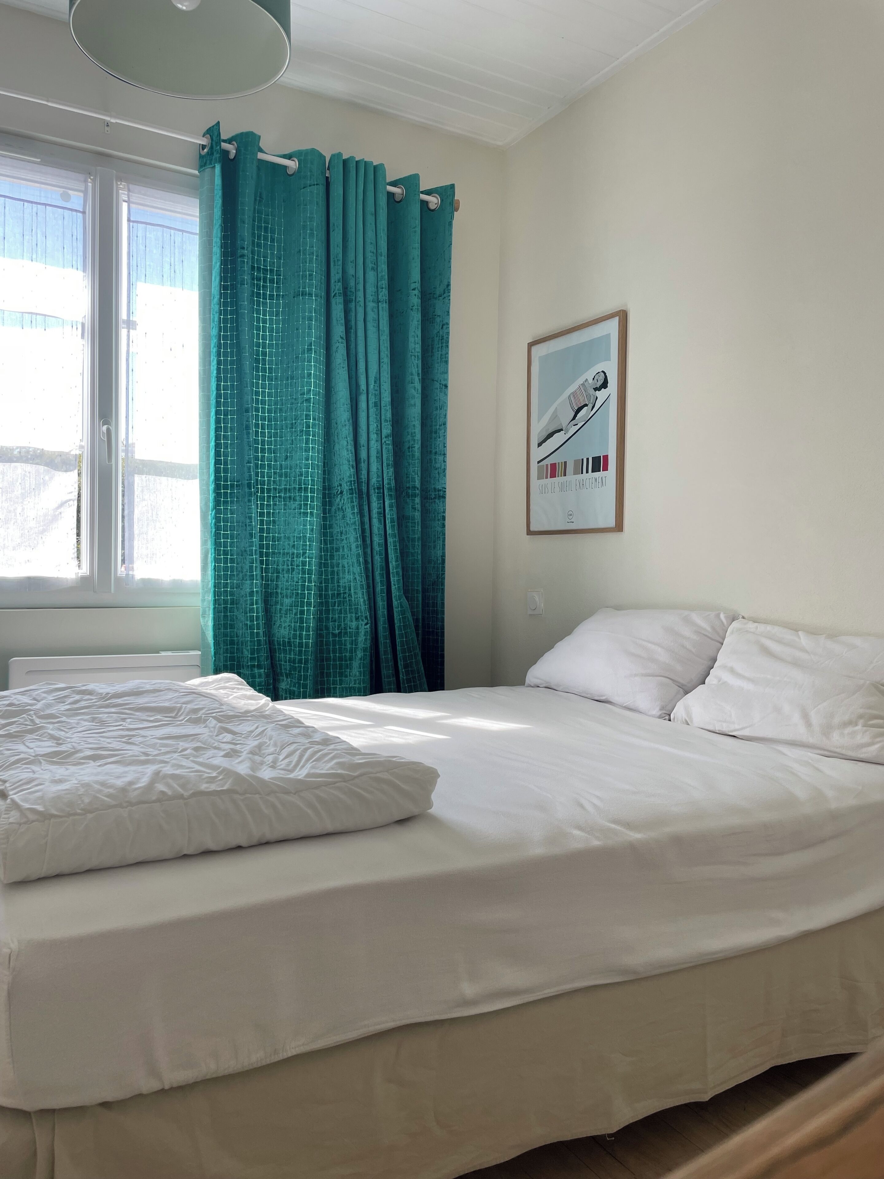 Een slaapkamer met een bed, gordijnen, een raam en een aan de wand bevestigd schilderij.