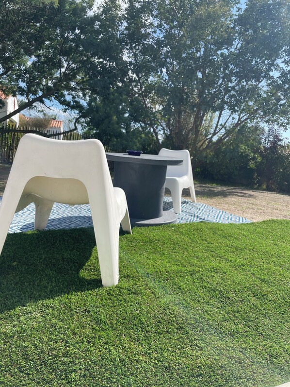 A patio area with white plastic chairs and a table, surrounded by artificial grass and trees.