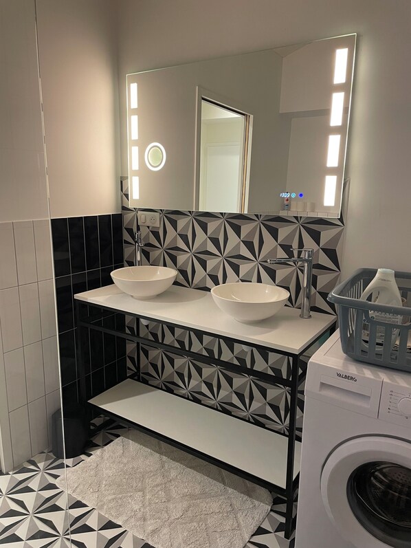 Un baño moderno con un lavamanos doble, un espejo grande, una lavadora y azulejos estampados.