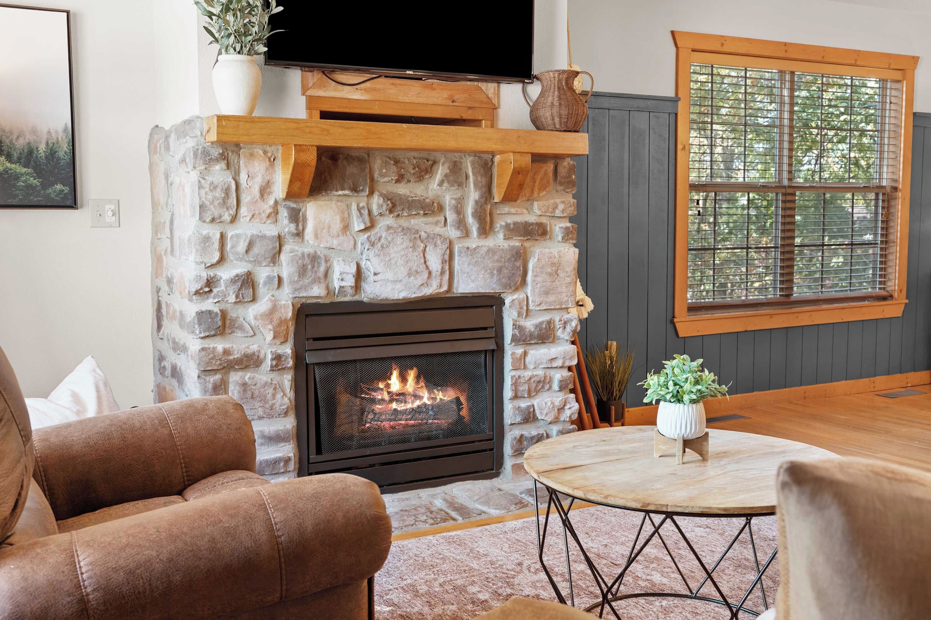 A living room with a stone fireplace, a television mounted above it, and a window with blinds.