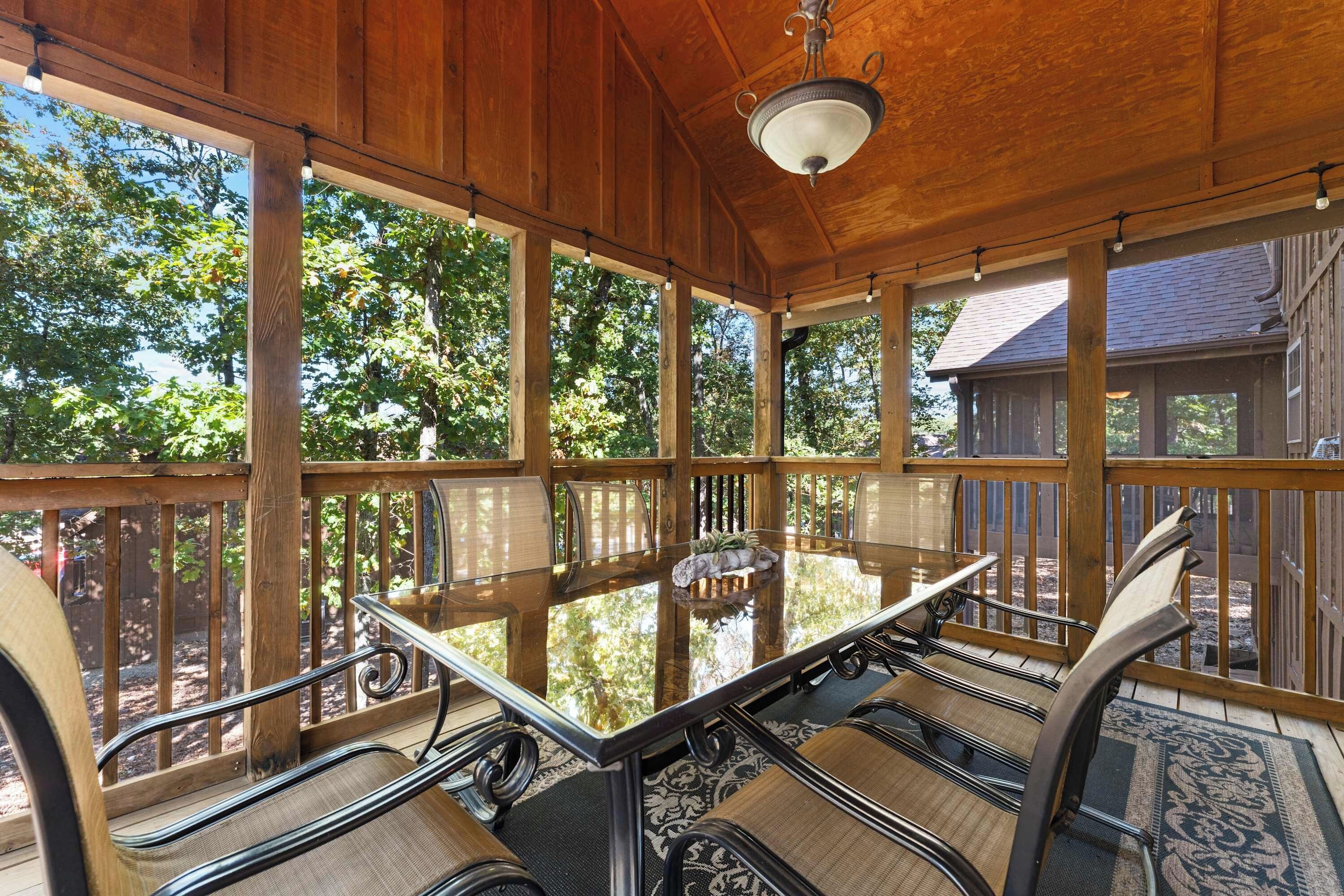 A covered patio with a glass-top table and metal chairs, surrounded by wooden railings and a view of trees.