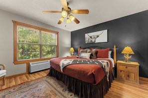 A bedroom with a large window, a ceiling fan, a bed with multiple pillows, a nightstand with lamps, and a wall-mounted TV.