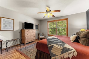 A bedroom with a ceiling fan, a television on a wooden dresser, a window with blinds, a patterned rug, and a bed with a throw blanket.