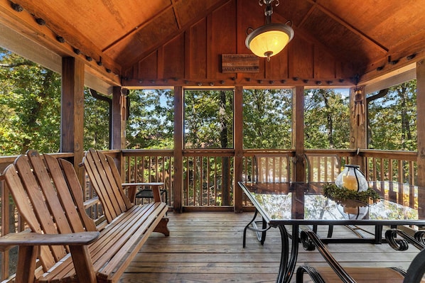 A wooden deck with benches and a table, surrounded by glass railings, offering a view of trees.