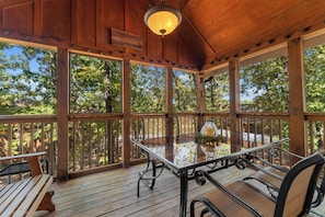 An outdoor wooden deck with a glass table and chairs, surrounded by a wooden railing and trees.