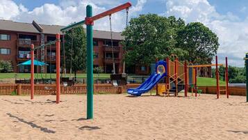 A playground with swings, a slide, and climbing structures, surrounded by a sandy area with a building in the background.