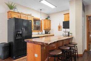 Una cocina con un refrigerador negro, gabinetes de madera, una isla de madera con taburetes y una ventana con persianas.