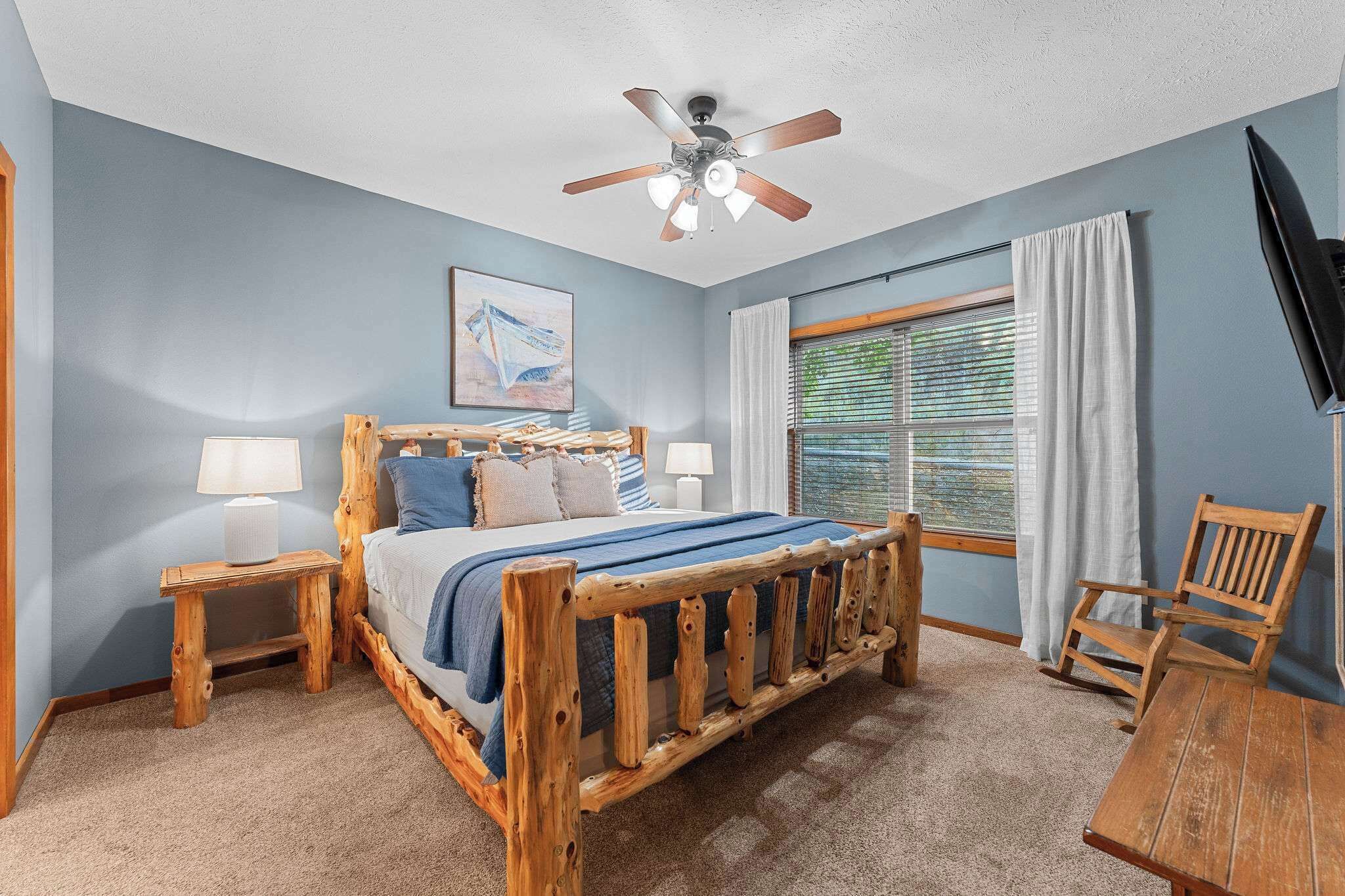 A bedroom with a wooden bed, two bedside tables, a ceiling fan, a window with blinds, a chair, and a TV.