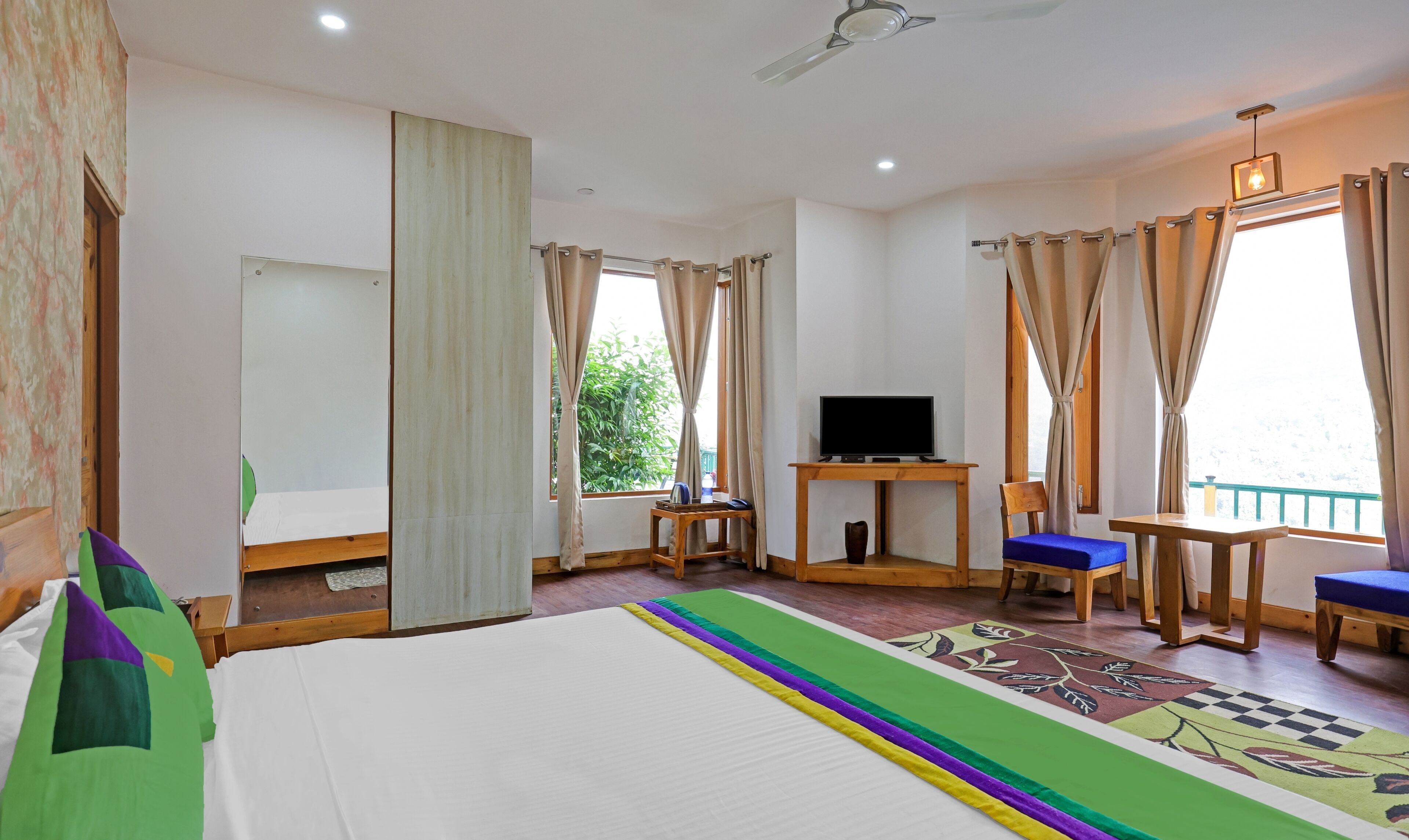A hotel room with a bed, a desk with a TV, a chair, and a view of greenery outside.