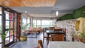 A restaurant with wooden ceiling, large windows, and brick walls. Tables and chairs are arranged for dining. - Treebo Vista Lago With Lake View (Nainital)