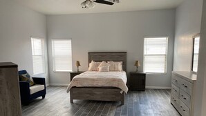 Un dormitorio con una cama, dos mesitas de noche, un tocador, una silla y dos ventanas con persianas.