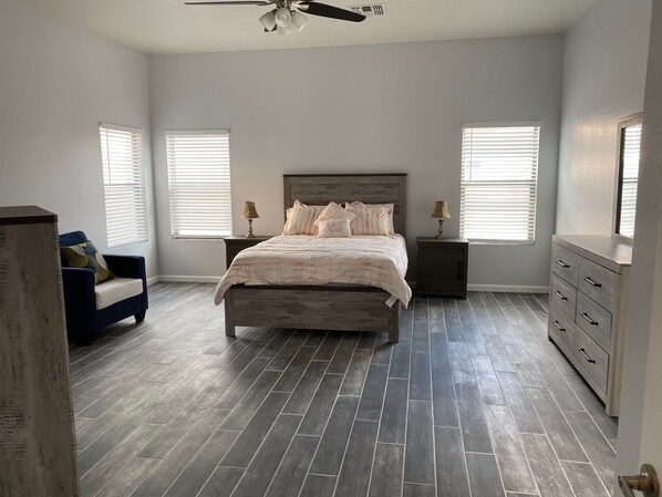 A bedroom with a bed, two nightstands, a dresser, a chair, and two windows with blinds.