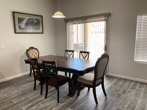 A dining area with a dark wooden table and six chairs, a pendant light, a framed picture on the wall, and a window with blinds.