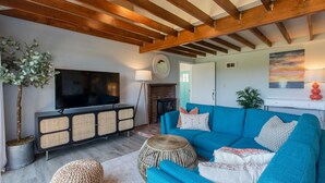 Ein Wohnzimmer mit einem großen Flachbildfernseher, einem blauen Sofa, einem Korbuscheinmöbel, einem runden Holzschemel und einem Kamin.