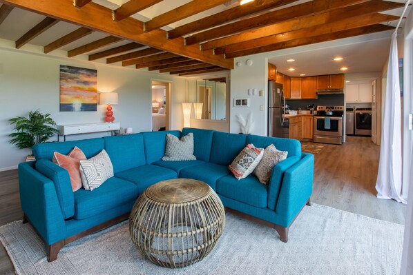 A modern living room with a teal sofa, a woven coffee table, and a kitchen in the background.