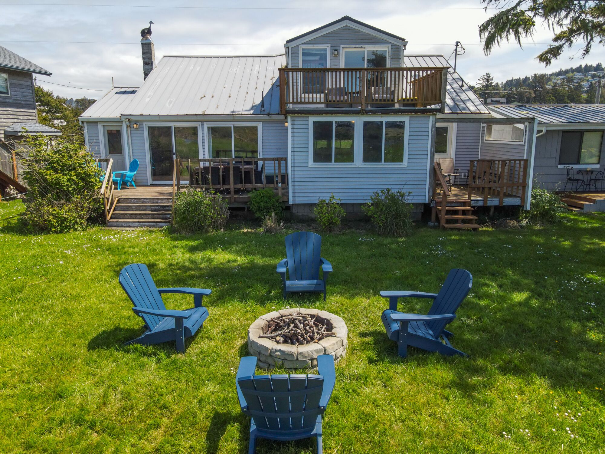 A house with a deck, a fire pit, and four Adirondack chairs in the yard.