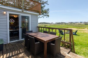 A wooden deck with a table and chairs, a glass door leading to a room, and a view of a grassy field and houses in the distance.