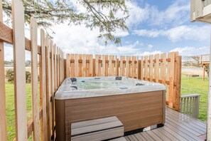 A hot tub surrounded by a wooden deck and fence, with a grassy yard and a house in the background.
