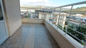 Un balcón con suelo de baldosas, barandilla metálica y vista a una zona residencial con edificios y colinas al fondo.