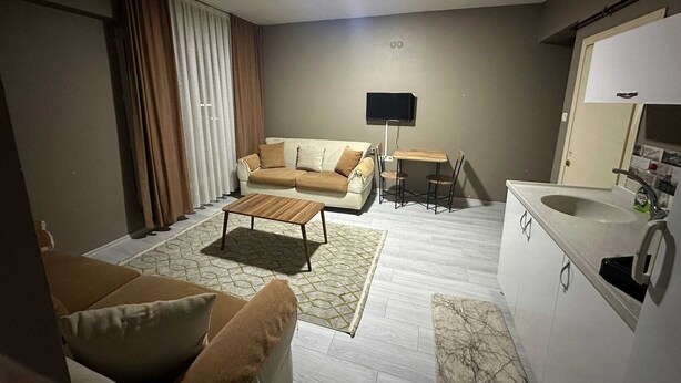 Una sala de estar moderna con un sofá, una mesa de centro y un televisor. Hay una zona de cocina con un lavamanos y armarios.