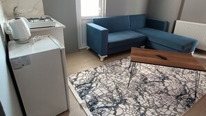 Una sala moderna con un sofá azul, una mesa de centro de madera, un tapete estampado, un refrigerador blanco y una zona de cocina con estufa y hervidor.