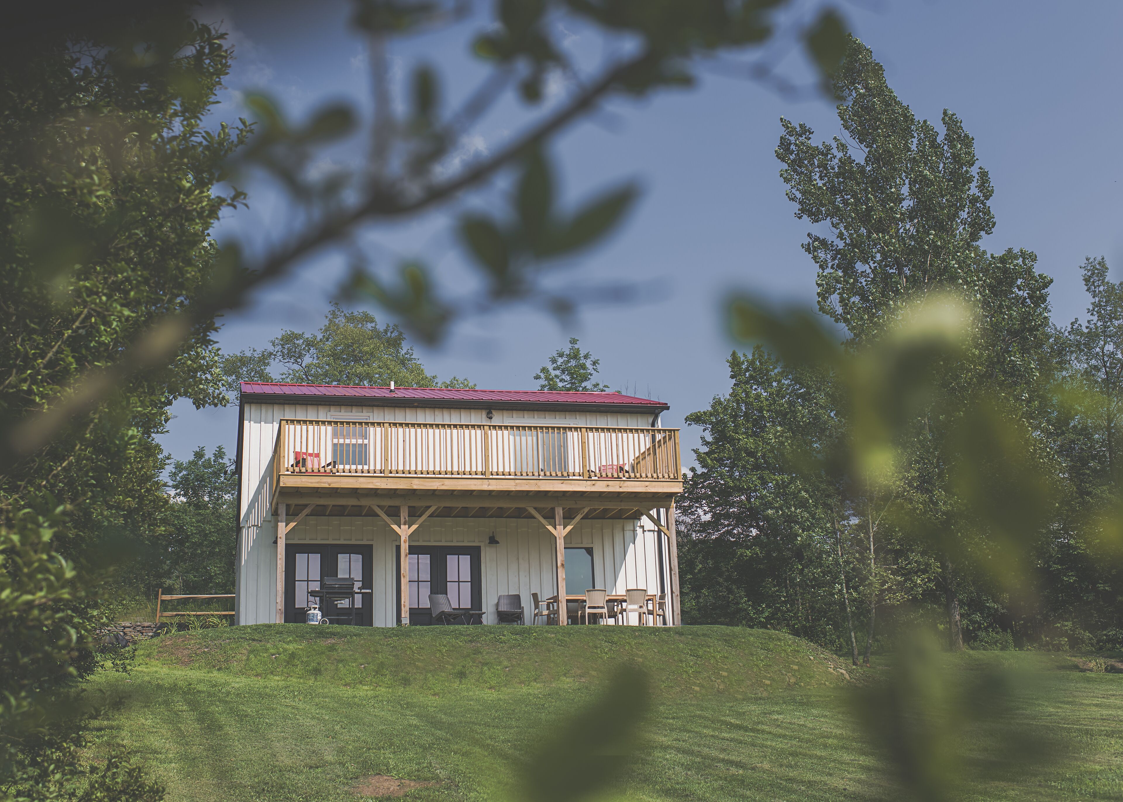 A two-story wooden house with a balcony, surrounded by greenery.
