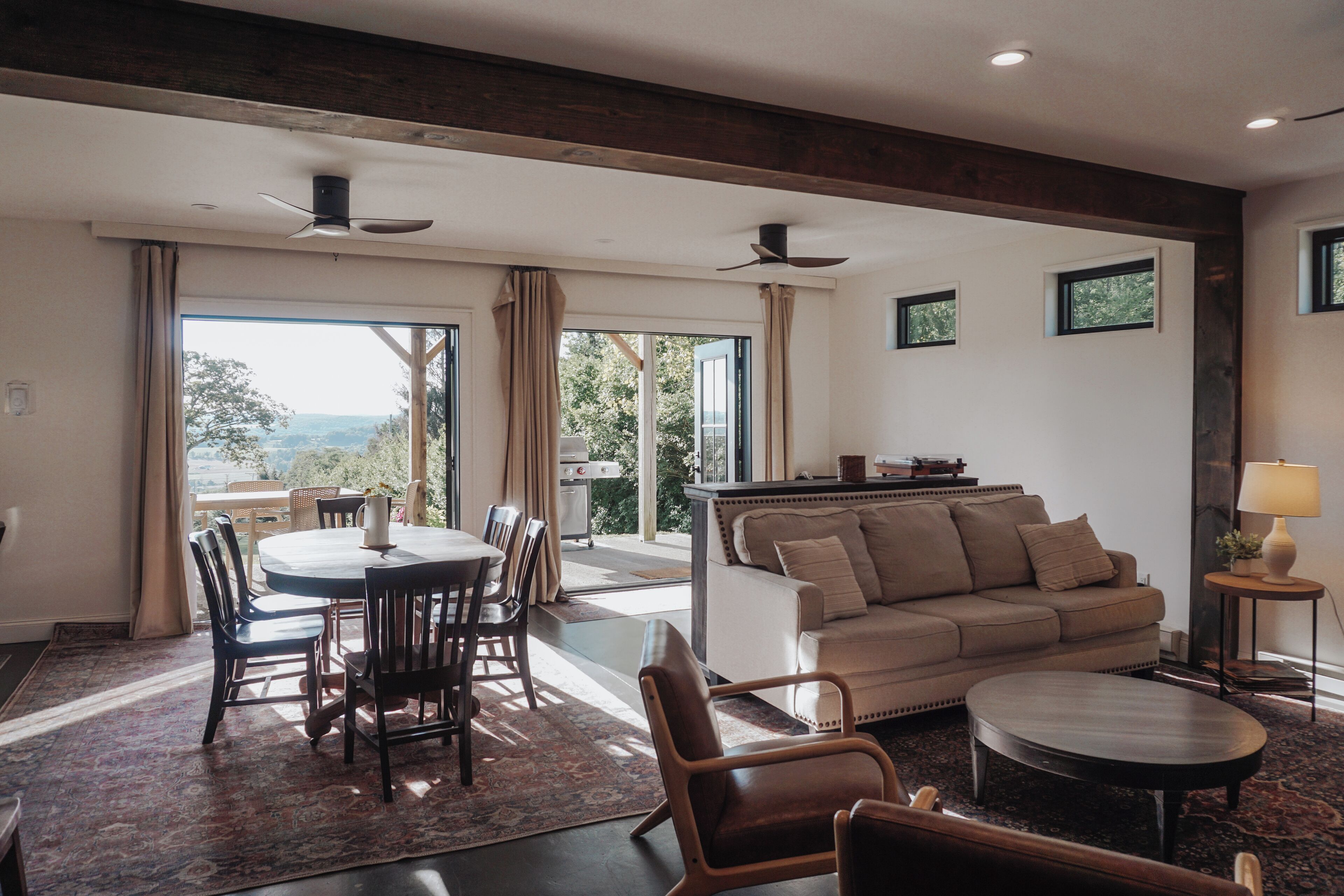 A spacious living area with a dining table, chairs, a sofa, and a coffee table. There are ceiling fans, a view through large windows, and a small side table with a lamp.