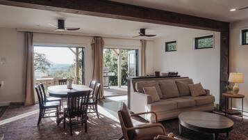 A spacious living area with a dining table, chairs, a sofa, and a coffee table. There are ceiling fans, a view through large windows, and a small side table with a lamp.