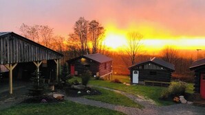A rustic cabin area with a covered structure and a small building with a red door, surrounded by trees and a sunset in the background. - Spacious Winter Retreat w/ Hot Tub, Games, Fire Pit & Scenic Hilltop Views! (Thompson)