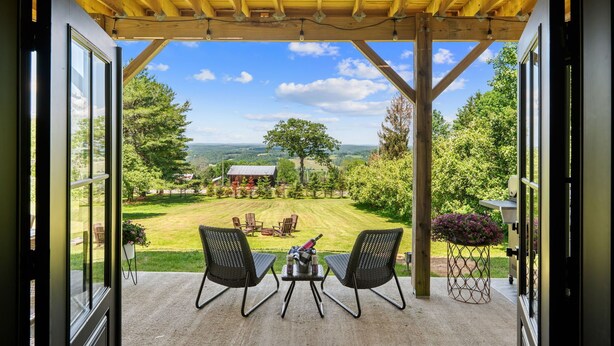An open patio with a wooden pergola, two chairs, a small table with a bottle and glasses, and a view of a grassy yard with trees and a house in the distance. - Spacious Winter Retreat w/ Hot Tub, Games, Fire Pit & Scenic Hilltop Views! (Thompson)