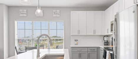 Una cocina moderna con armarios blancos, una isla grande con un lavamanos, un refrigerador de acero inoxidable y vistas a un estacionamiento a través del ventanal.