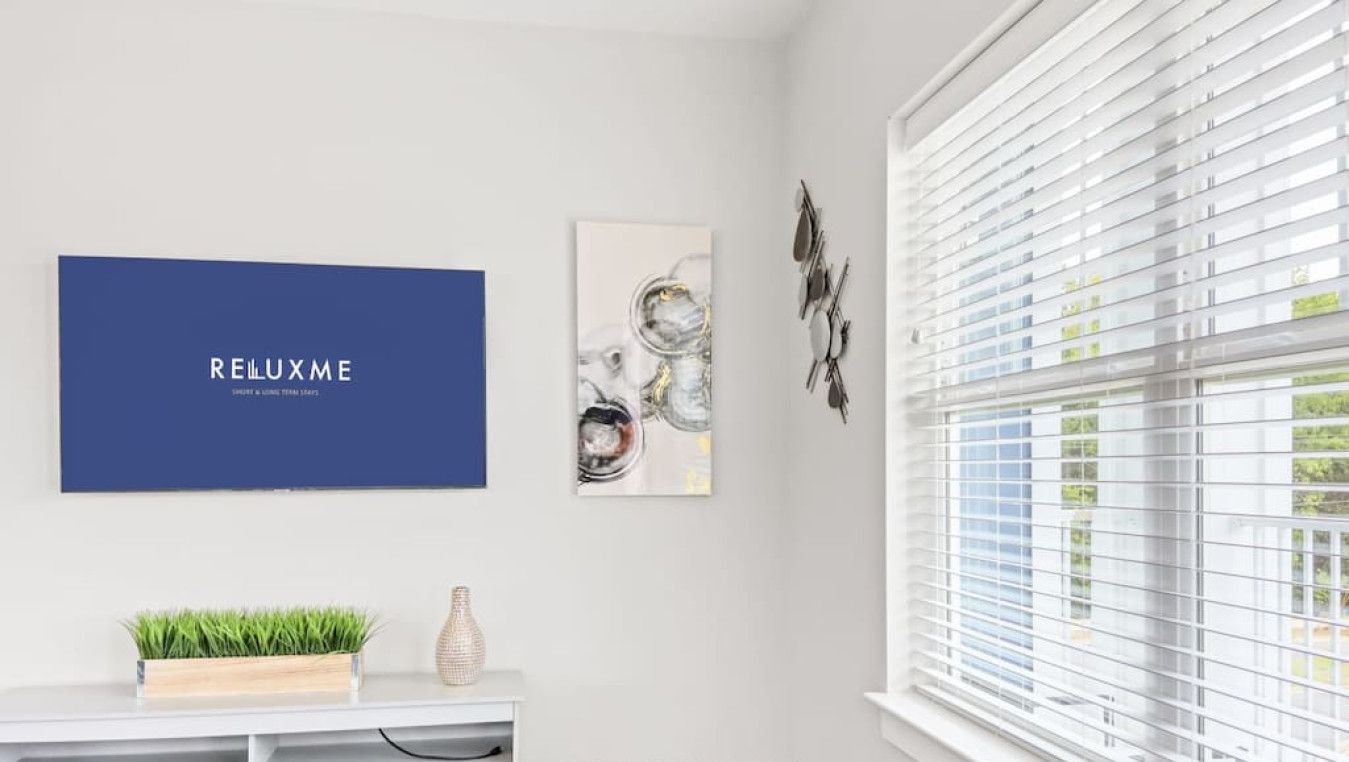 A room with a white wall, a blue framed artwork with the text "REILUXME", a small white console table with a plant and a vase, and a window with blinds.