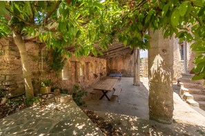 An outdoor patio area with a wooden table and benches, surrounded by stone walls and trees.