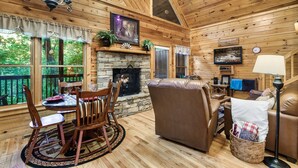 Ein gemütliches Blockhaus mit Kamin aus Stein, Holzwänden, einer Essecke mit Stühlen und Tisch, einem Ledersofa und einem an der Wand befestigten Fernseher.