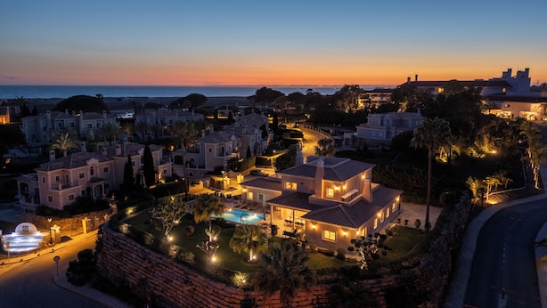 Een luxe villa met een zwembad, omgeven door een aangelegde tuin en uitzicht op zee bij zonsondergang.