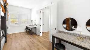 Una camera d'hotel moderna con pavimento in legno, una scrivania con sedia, uno specchio rotondo e una piccola area salotto con degli sgabelli.