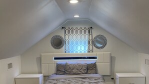 A bedroom with a sloped ceiling, a bed with a mattress and pillows, two bedside tables with drawers, a window with patterned curtains, and a wall-mounted mirror. - beautiful Modern apartment, close to everything ! (Hicksville)