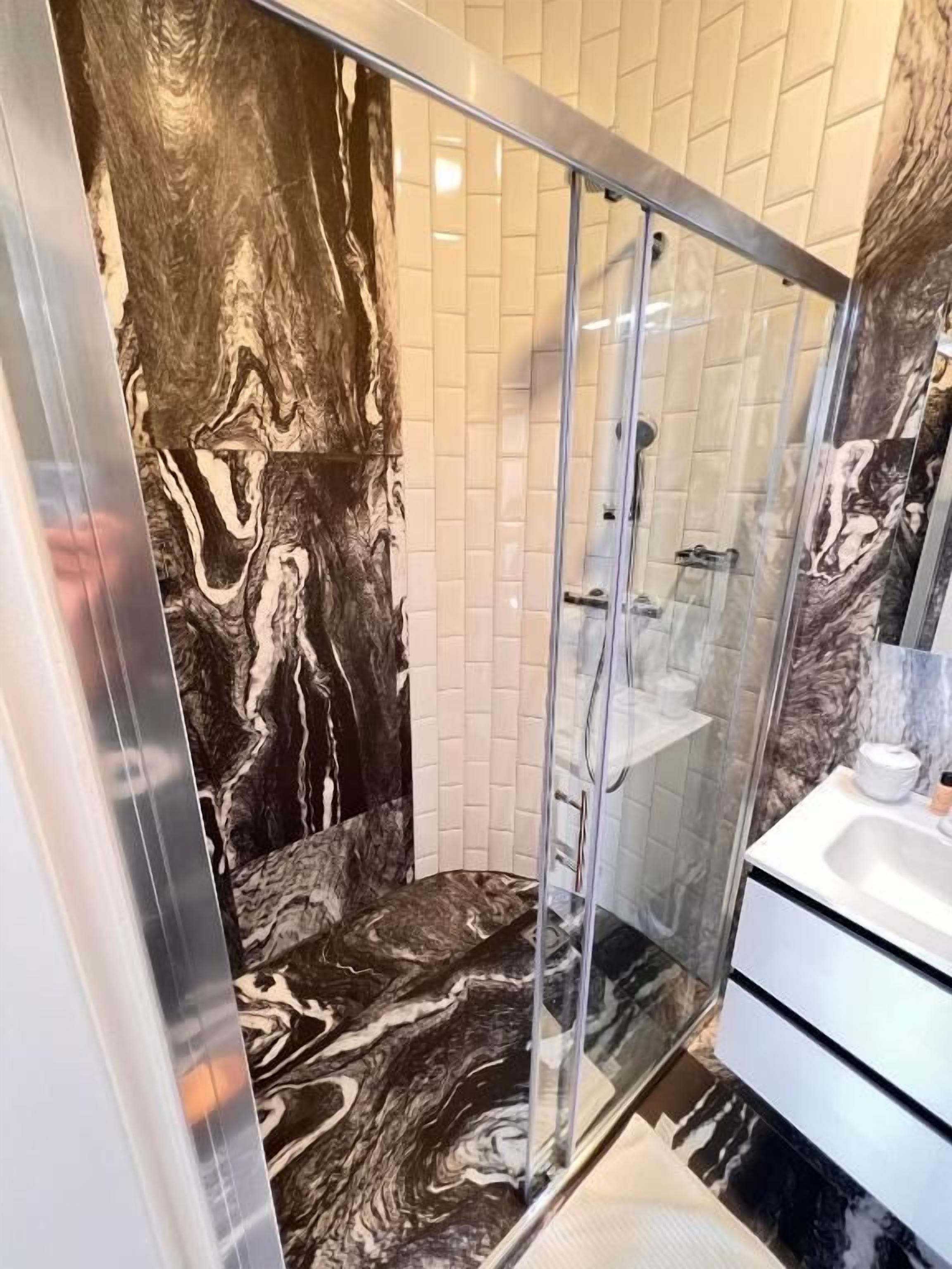 A modern bathroom with a marble shower wall, a glass-enclosed shower, and a white sink with a marble countertop.