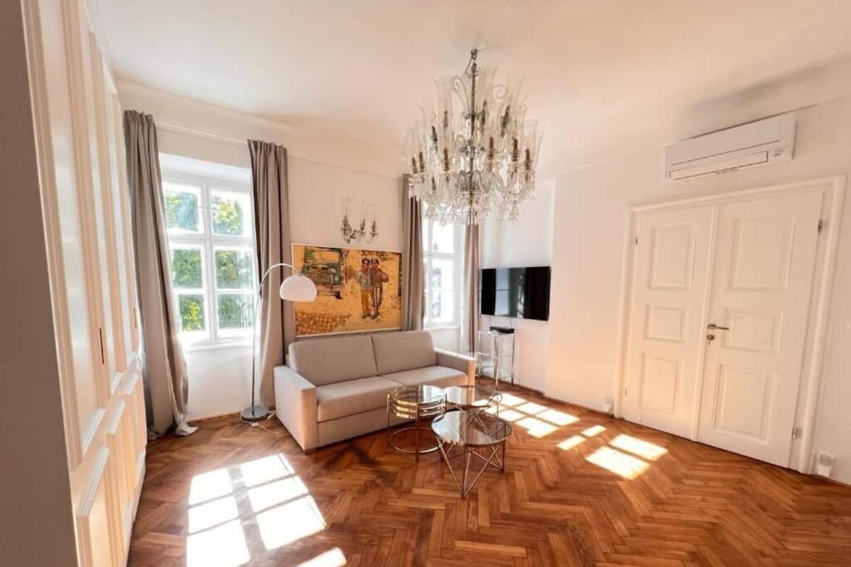 A spacious living room with a herringbone wooden floor, a chandelier, a sofa, and a painting on the wall.