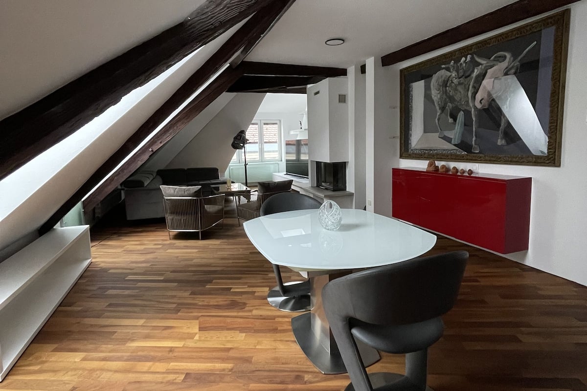 A modern dining area with a white table and black chairs, wooden flooring, and a sloped ceiling with exposed beams.